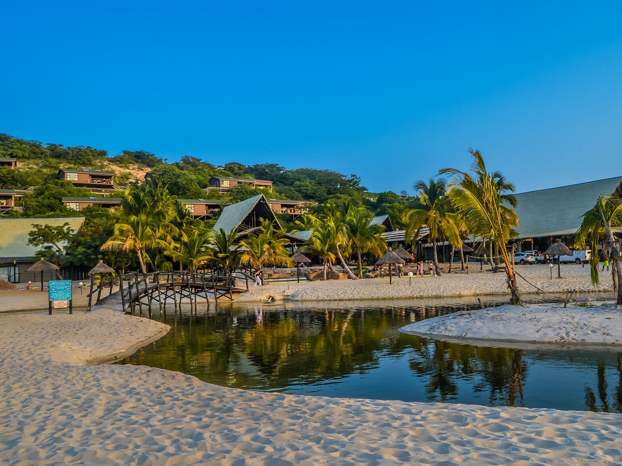 Playa en Maputo con un río que pasa por la arena, un puente para cruzar el pequeño río y palmeras dispersas
