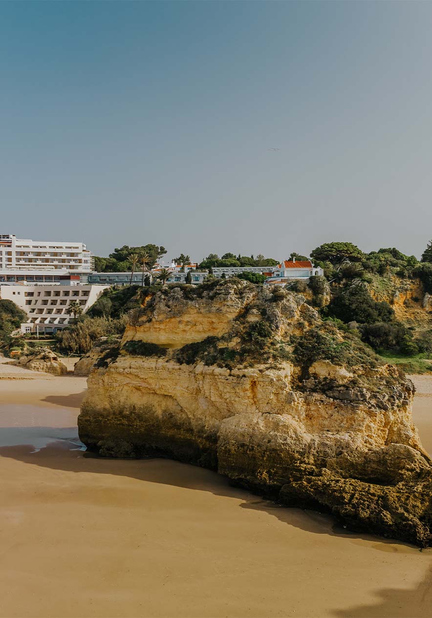 Playa de Alvor con rocas, arena, vegetación, acantilado y edificios al fondo, bajo cielo despejado