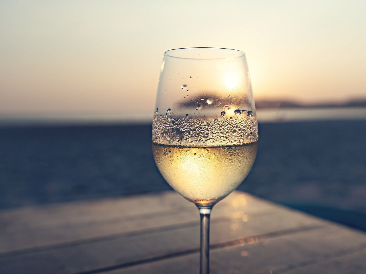 Copa de vino blanco con reflejos desenfocados del atardecer al fondo, sobre una superficie de madera