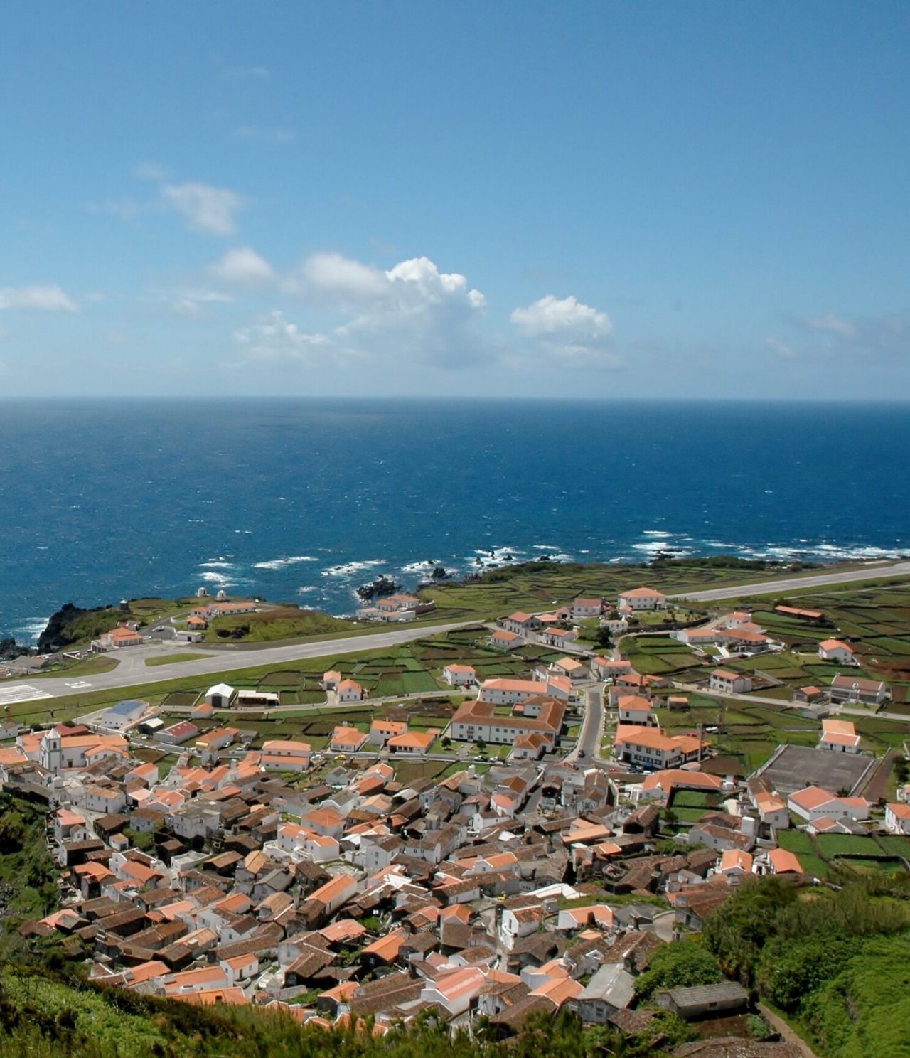 Vista aérea da ilha do Corvo, com a sua pista de avião, vila pitoresca e piscina natural, rodeada pelo mar azul profundo