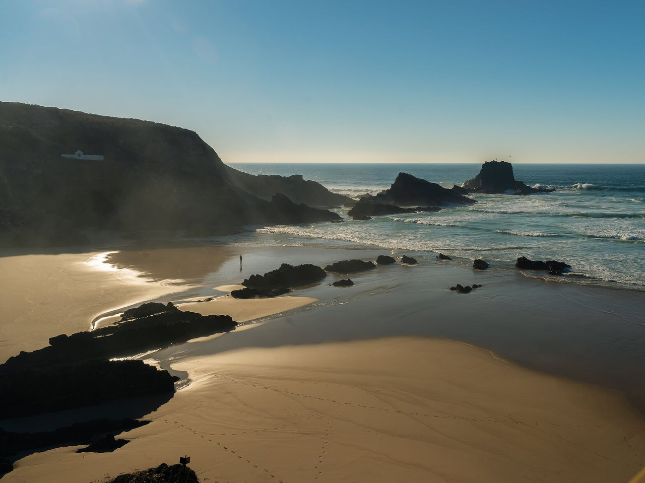 Vista aérea de uma praia deserta com falésias rochosas, mar agitado, e uma pessoa a caminhar sob um céu azul
