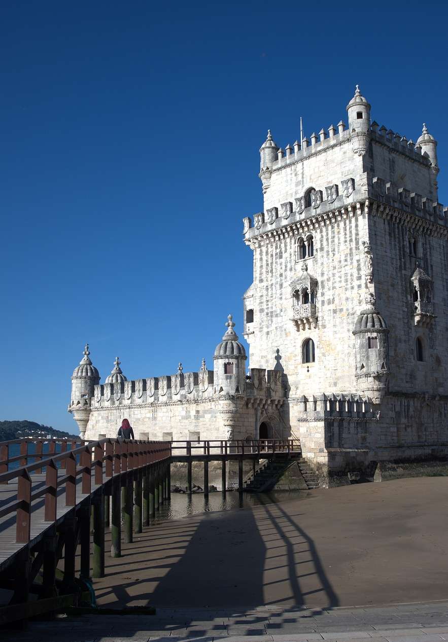 Torre de Belém com o passadiço de madeira na lateral, areia escura, o Rio Tejo por trás da Torre e árvores em redor
