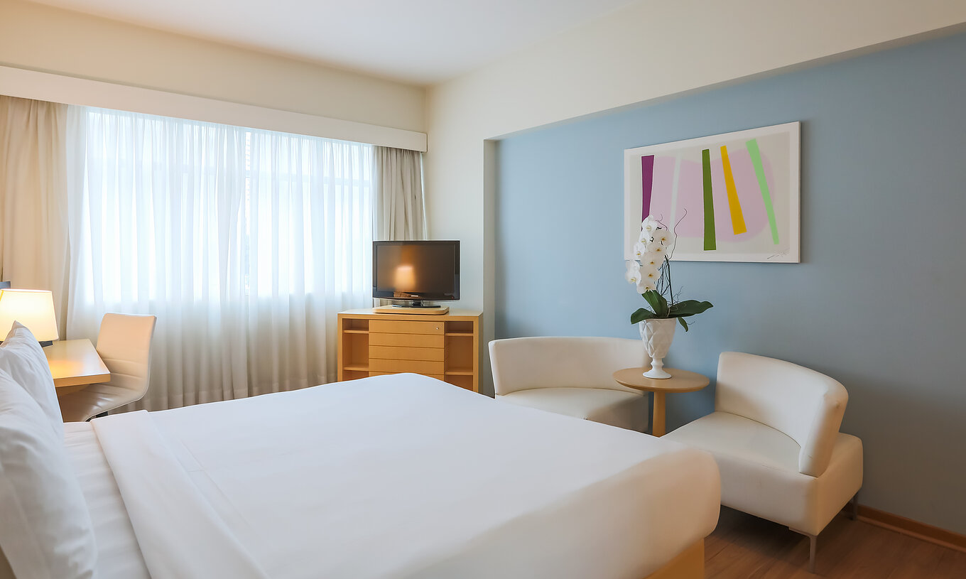 The Deluxe Corner at Pestana Curitiba has a double bed, two sofas, a furniture unit with a TV, and a desk