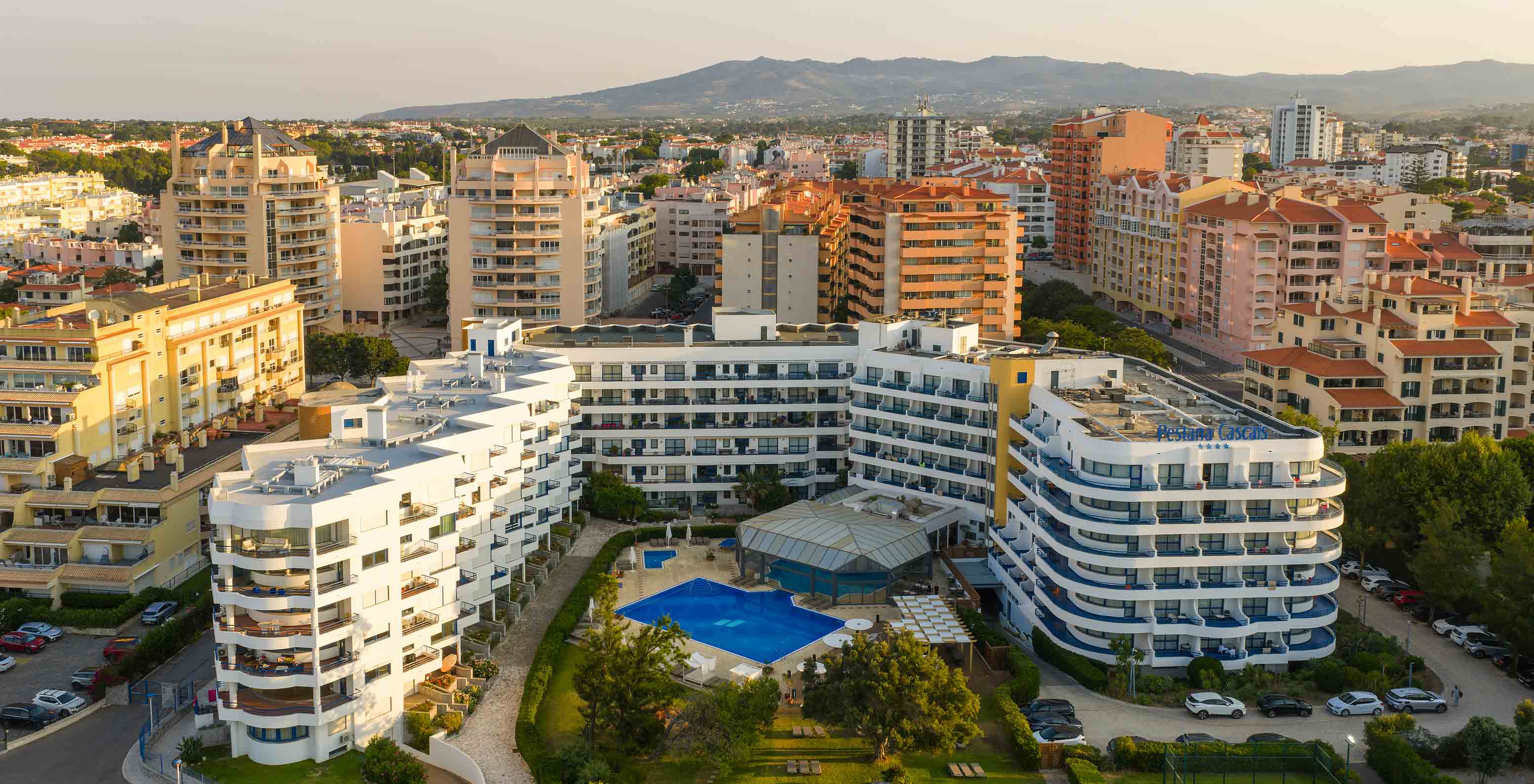 Aerial view of the hotel with balconies, pools, garden, and the city behind with mountains in the background