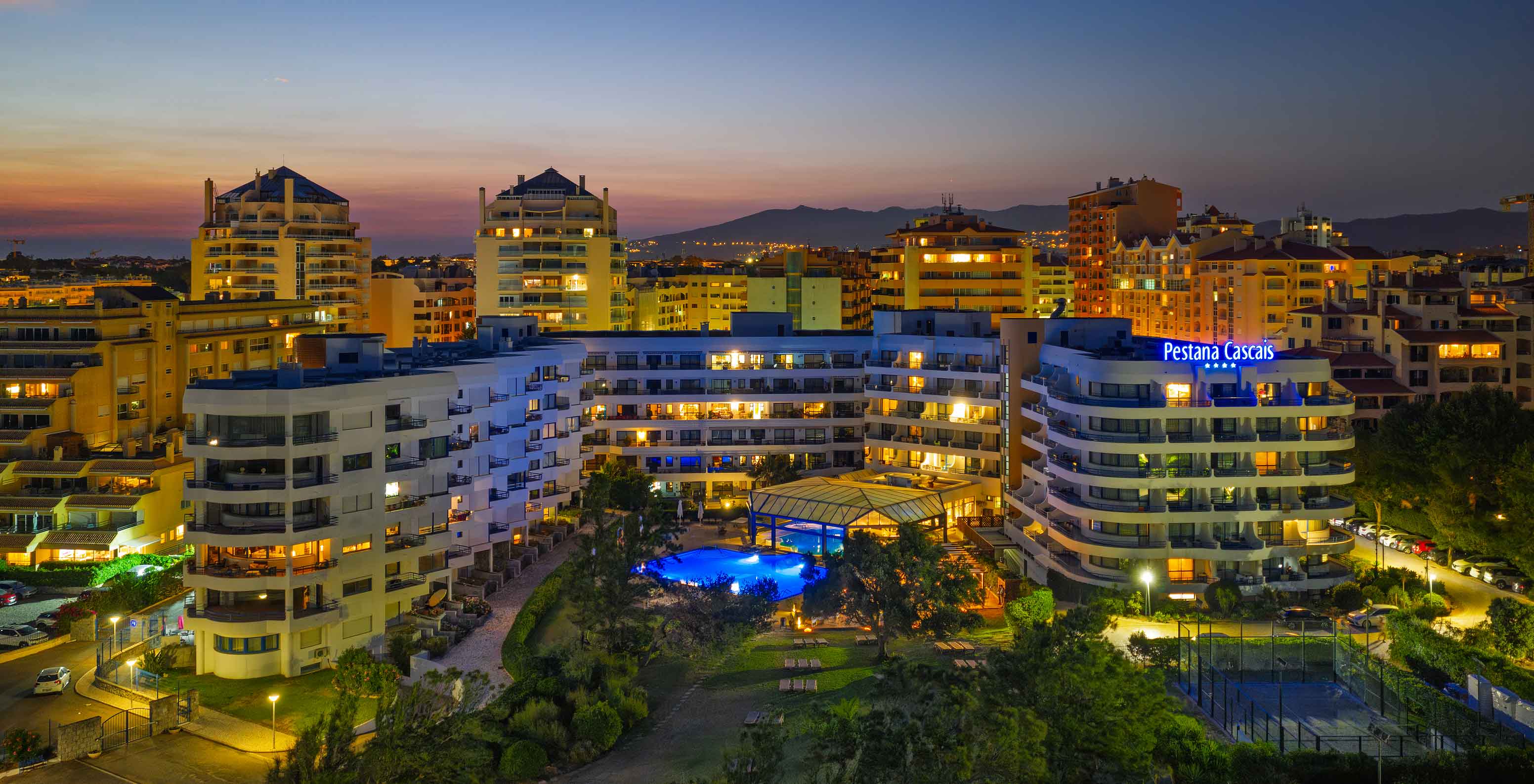 Aerial view of Pestana Cascais at sunset, with the city behind and a pink-orange sky in the background