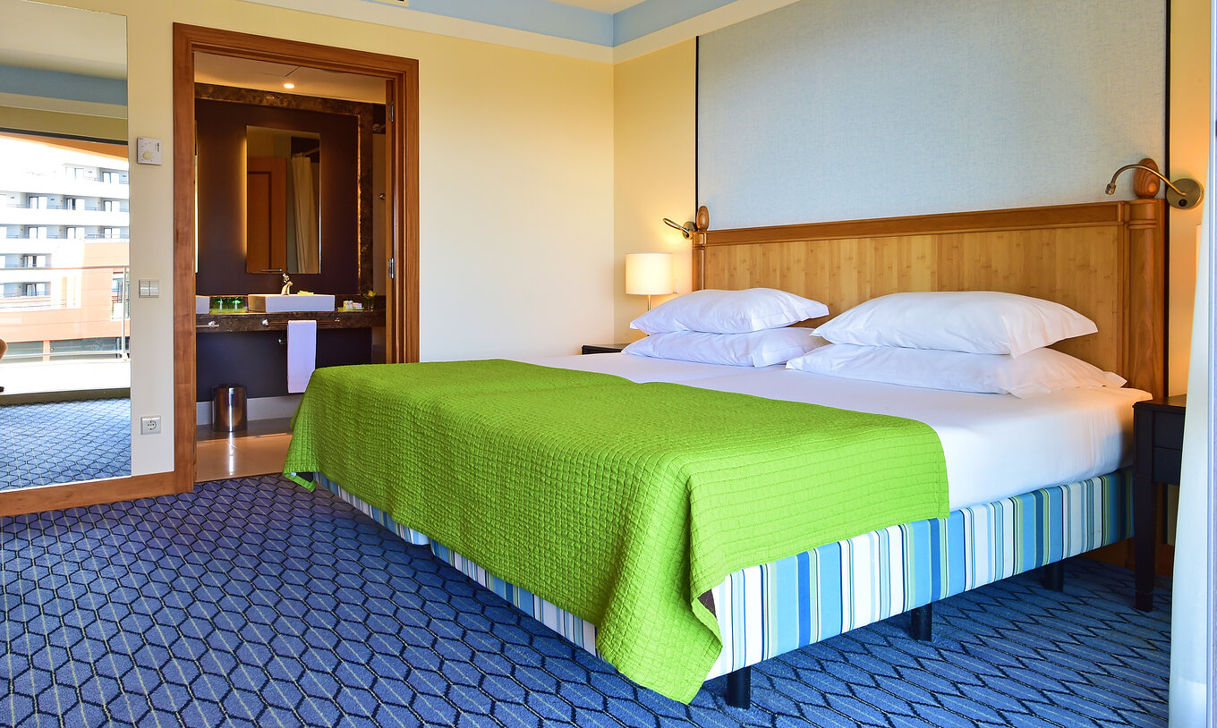 The Suite of Pestana Promenade has a double bed with a green bedspread, a tall mirror, and a bathroom