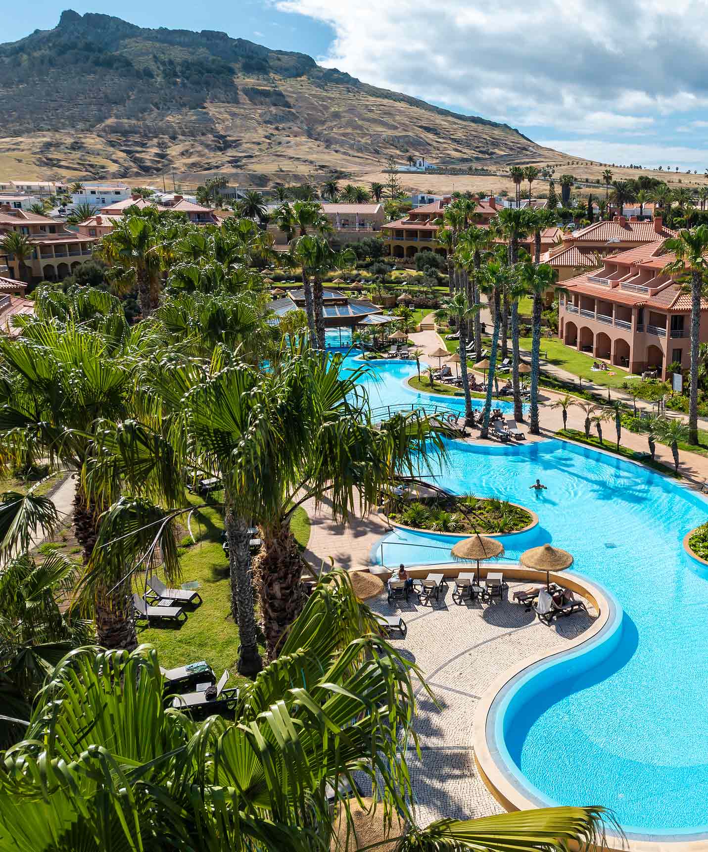 View of the outdoor pool at Pestana Porto Santo All Inclusive, surrounded by gardens, palm trees, and sun loungers.