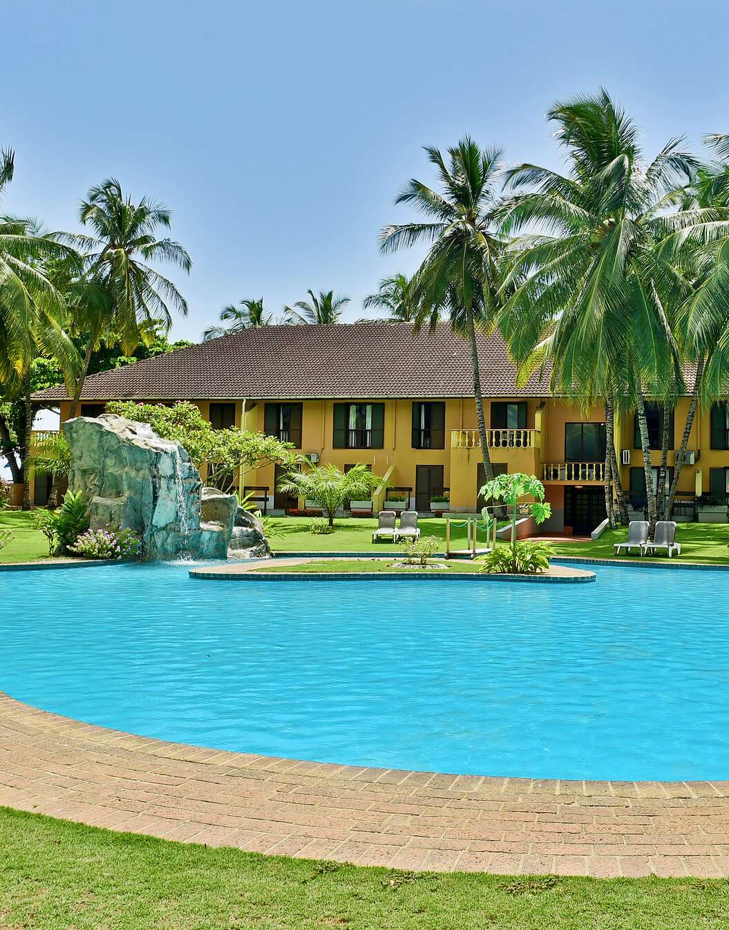 View of the yellow building with several windows, outdoor pool, palm trees, and sun loungers of a 4-star hotel in São Tomé