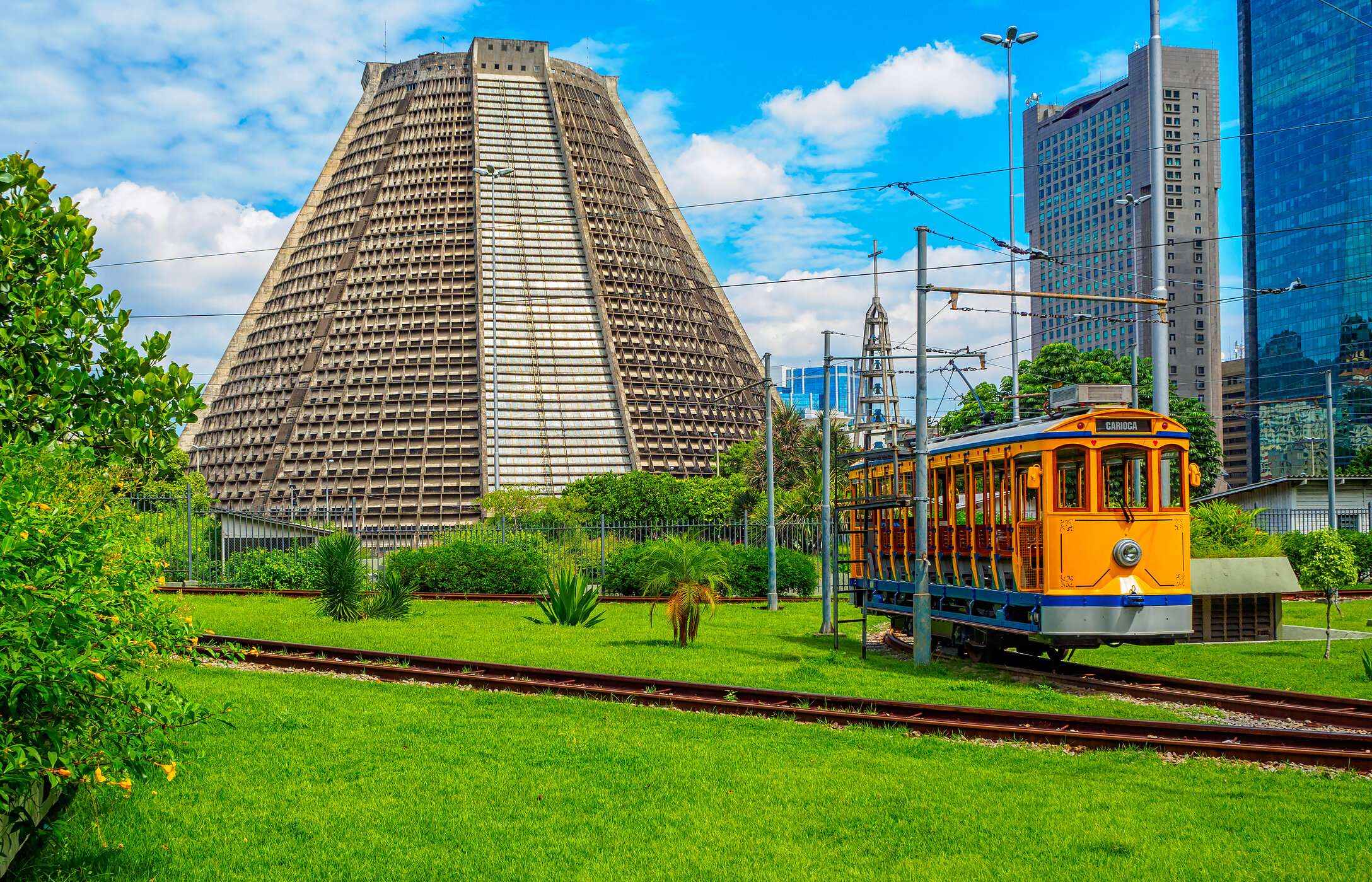 Catedral Metropolitana de Río de Janeiro, rodeada de vegetación, un tranvía antiguo y edificios al fondo.