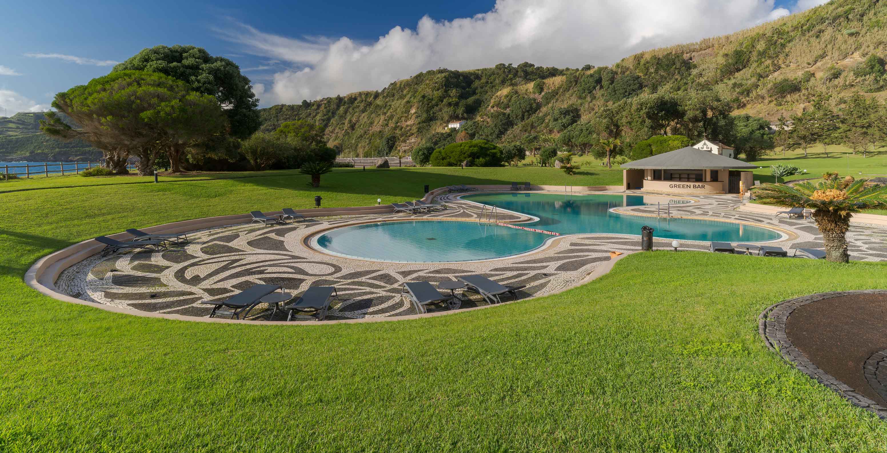 Piscina del Pestana Bahia Praia rodeada de jardín y colinas, con bar al fondo y cielo parcialmente nublado