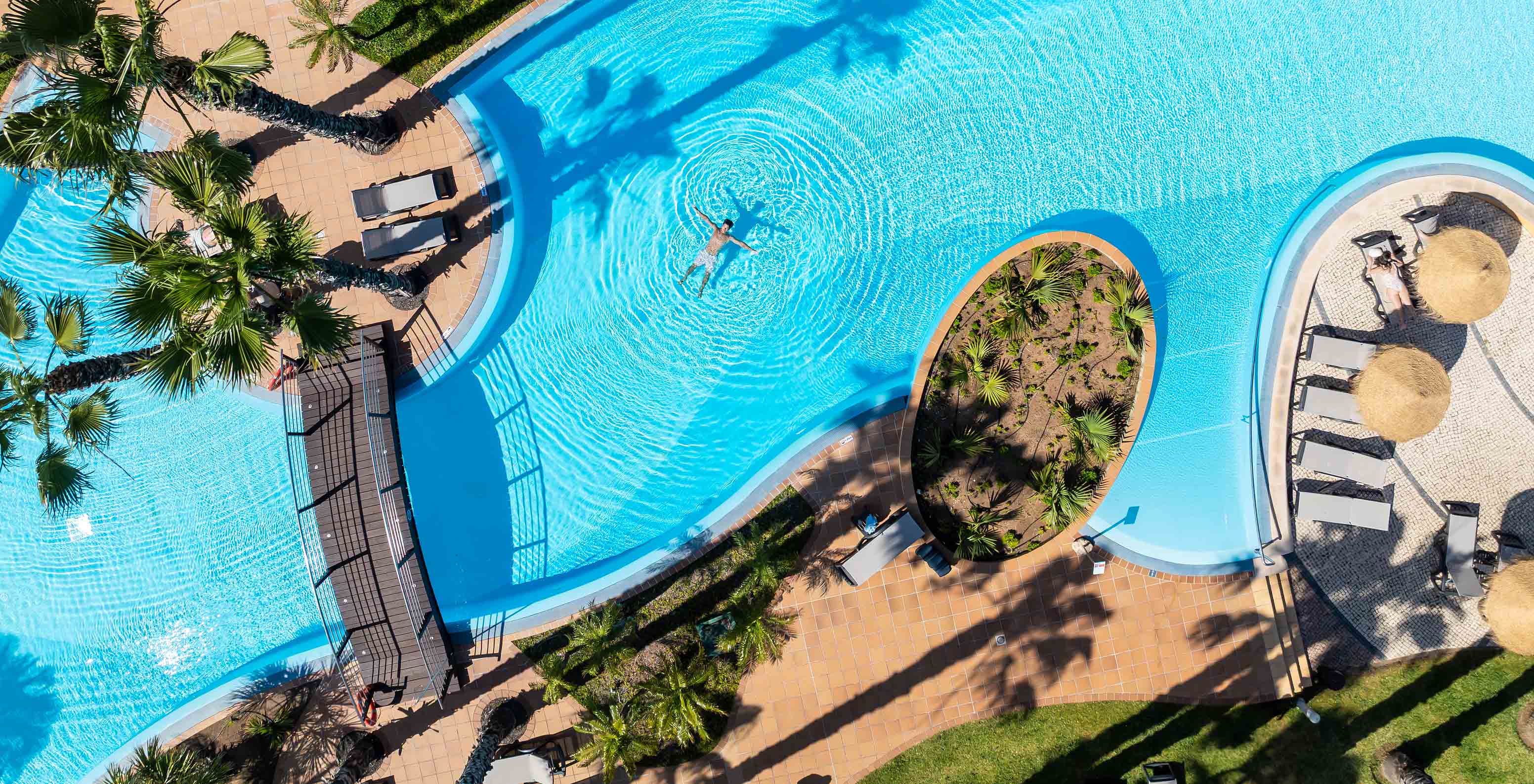 Vista de la piscina exterior del Pestana Porto Santo, con un huésped dentro del agua, un puente y palmeras tropicales