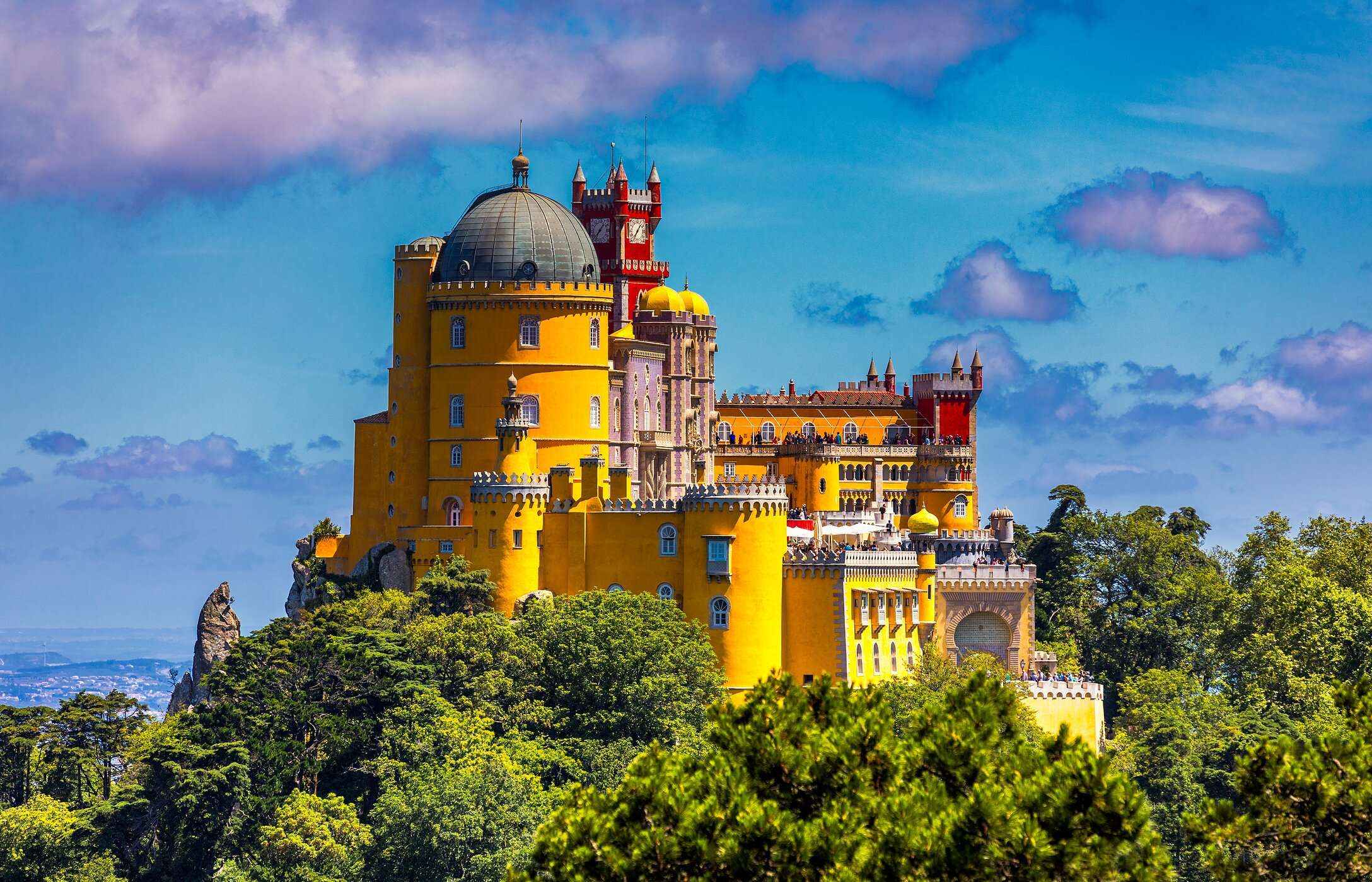 Vista aérea del Palacio da Pena en Sintra, con sus colores vibrantes, rodeado de vegetación bajo un cielo azul.