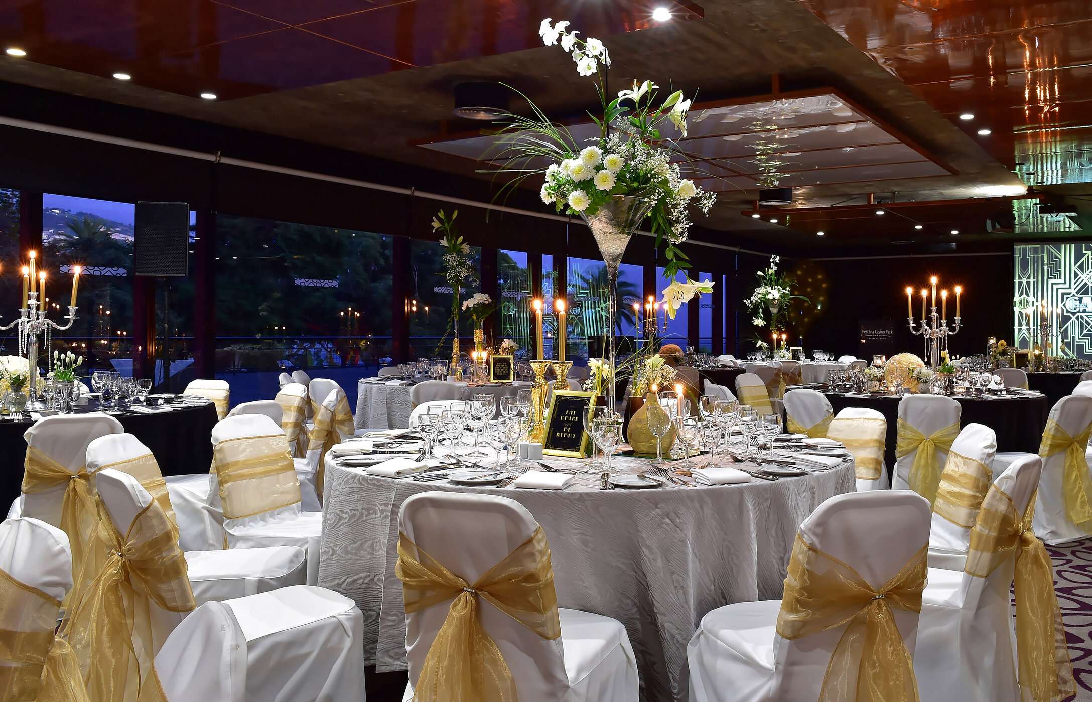 Boda en una sala con decoración moderna, mesas redondas con arreglos florales altos e iluminación suave y elegante