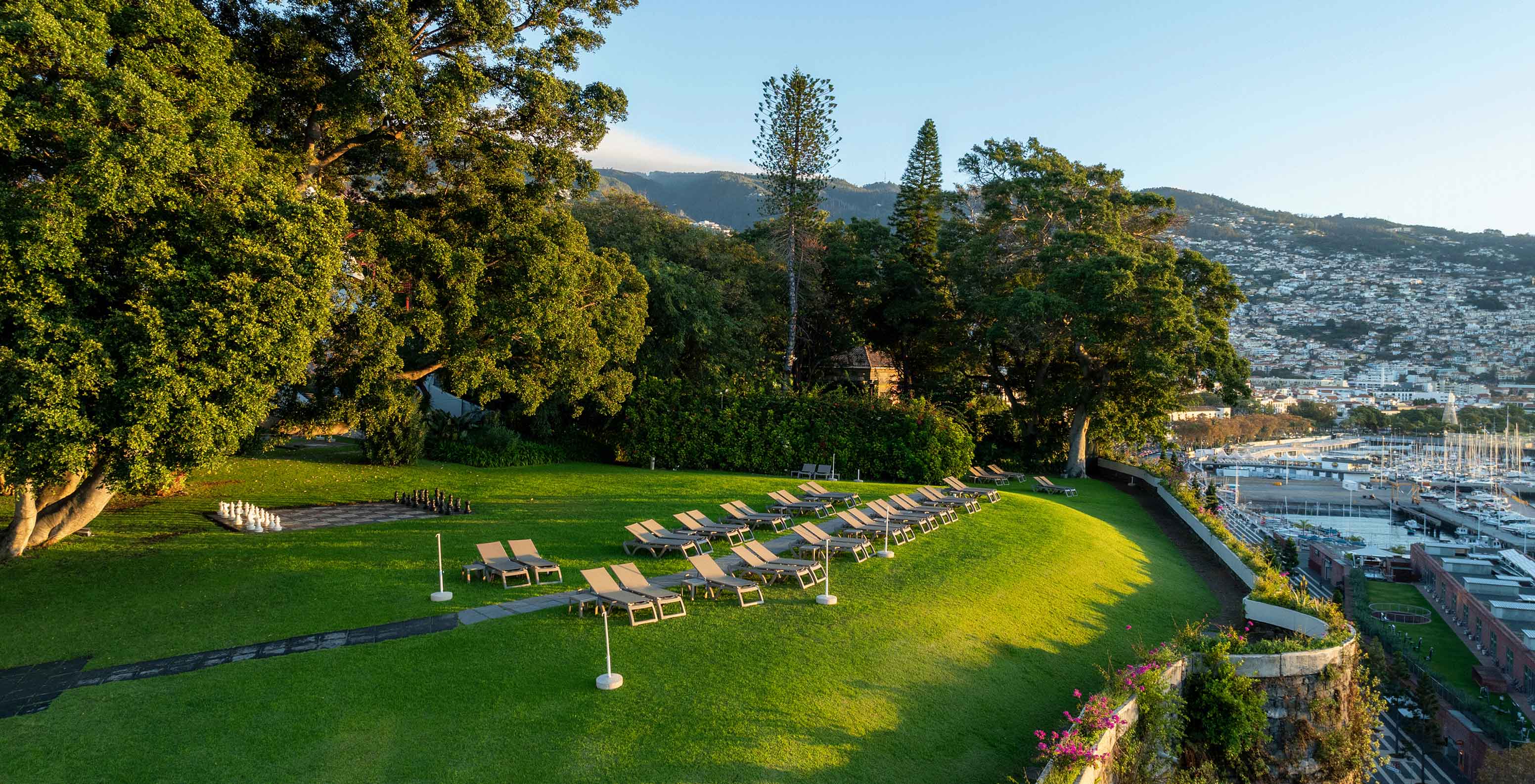 Uitzicht op de tuin van het Pestana Casino Park bij zonsondergang, met een stad op de achtergrond en een cruiseschip voor anker in de zee.