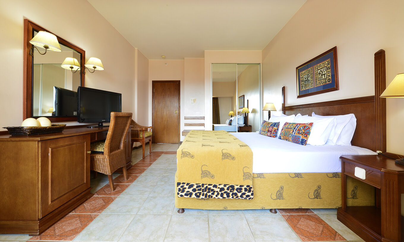 A Suite Luxo do Pestana Rovuma tem uma cama de casal e um quadro com decoração típica africana, uma secretária e televisão