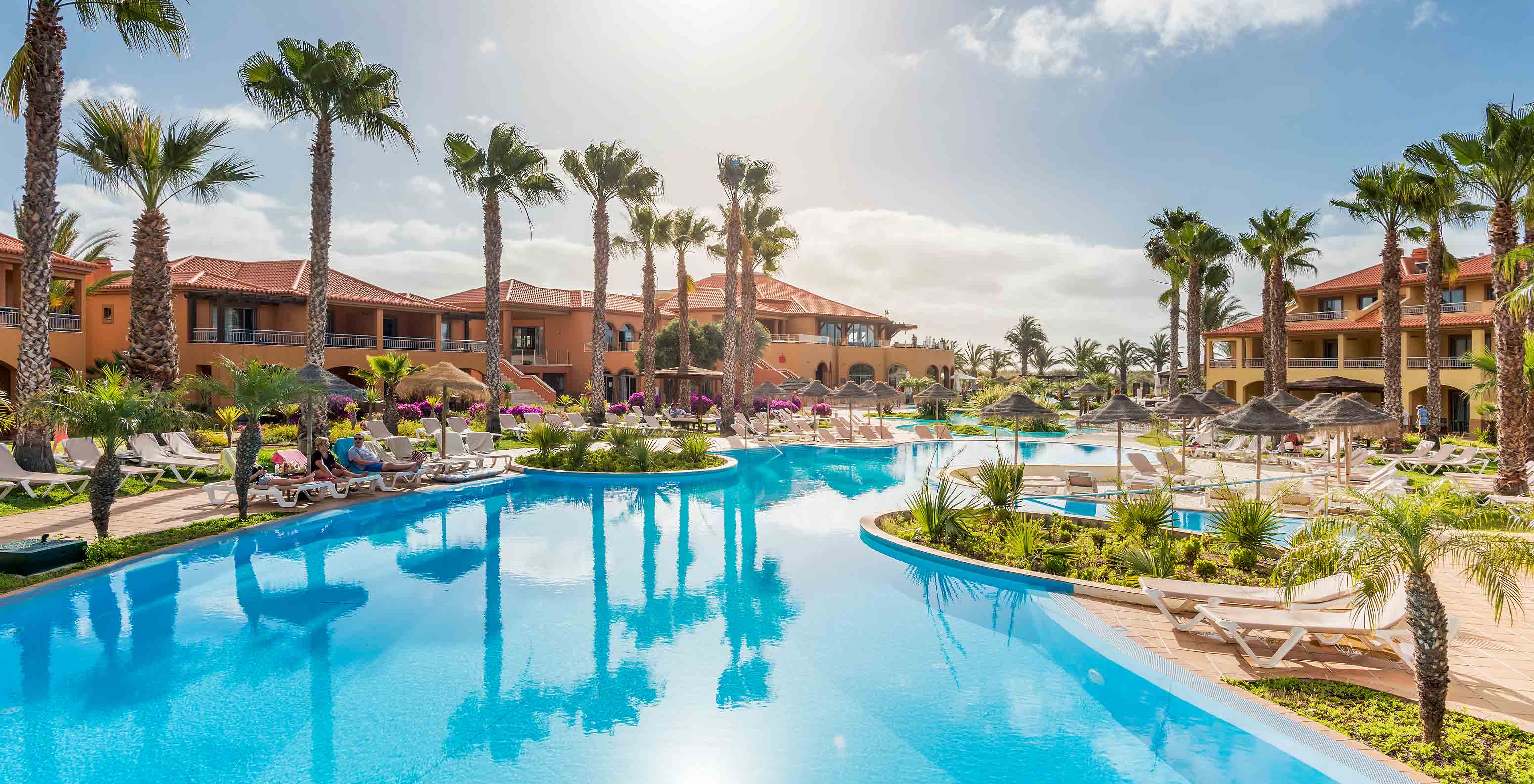 Ampla piscina azul do Pestana Porto Santo, rodeada por espreguiçadeiras, palmeiras e edifícios sob céu ensolarado