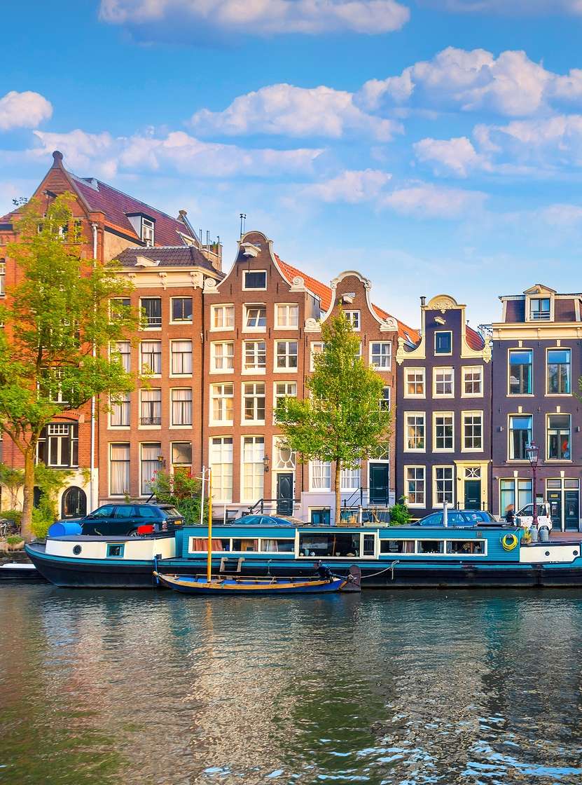 Canal de Amsterdão com pessoas a caminhar pela margem, barcos, e edifícios típicos, com um céu azul 