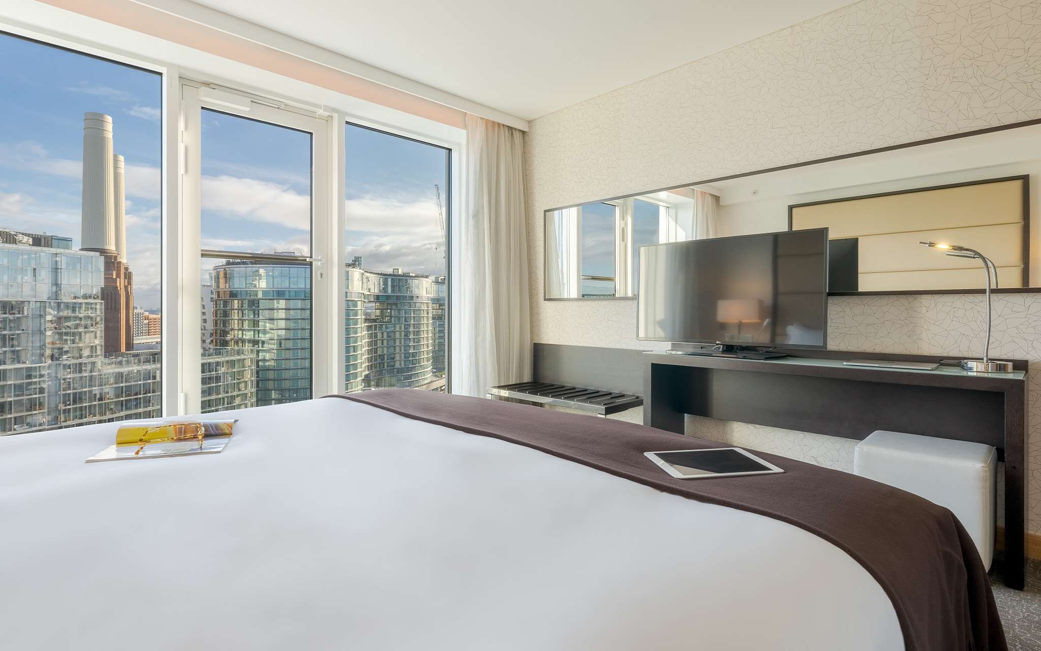 Quarto com cama de casal, televisão, espelho e janelas grandes com vista para a cidade de Londres