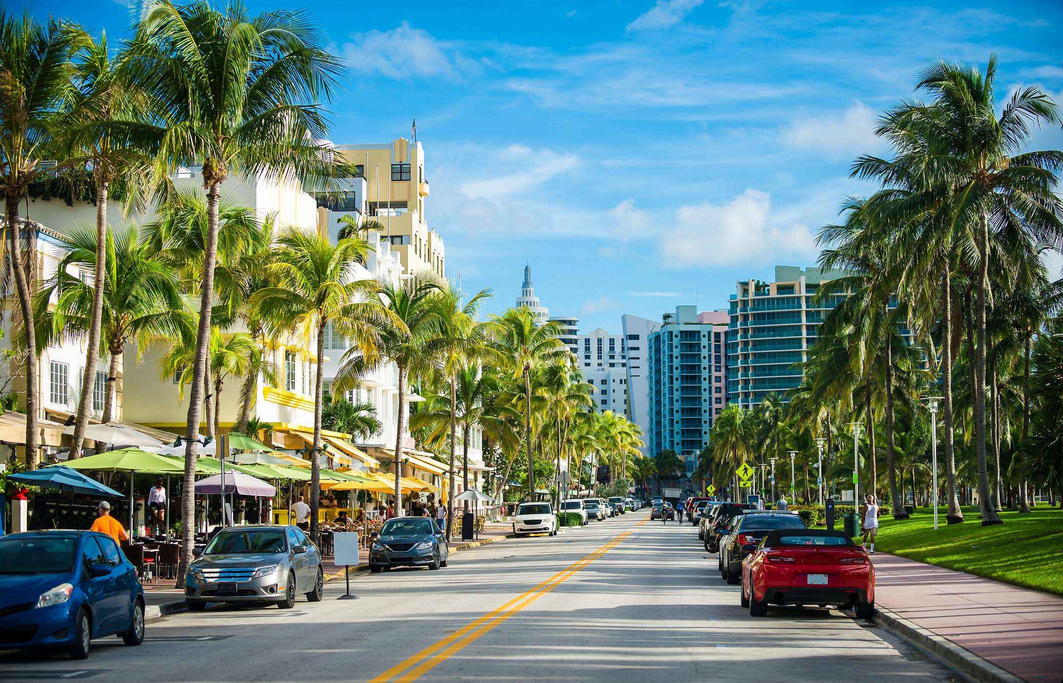 Vista sobre a Ocean Drive em Miami, com palmeiras altas, edifícios Art Déco e vários carros estacionados