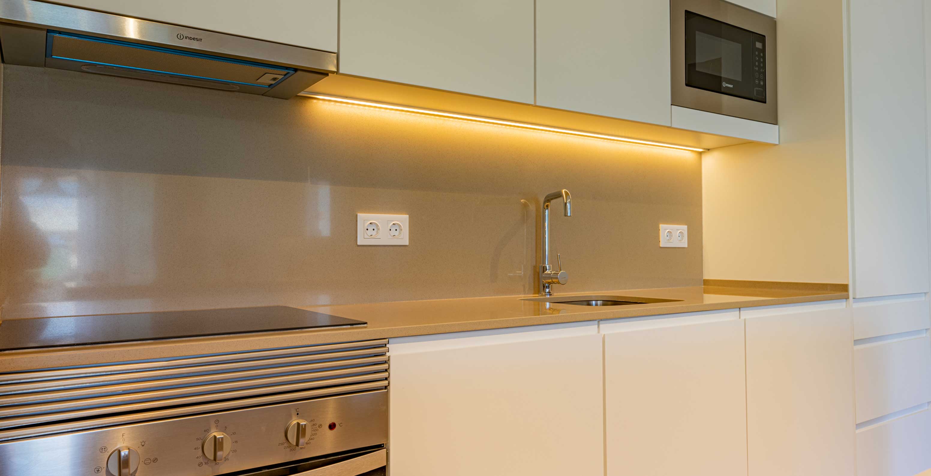 Cozinha moderna com bancada bege, placa elétrica, forno, micro-ondas e armários brancos no Pestana Porto Covo Village