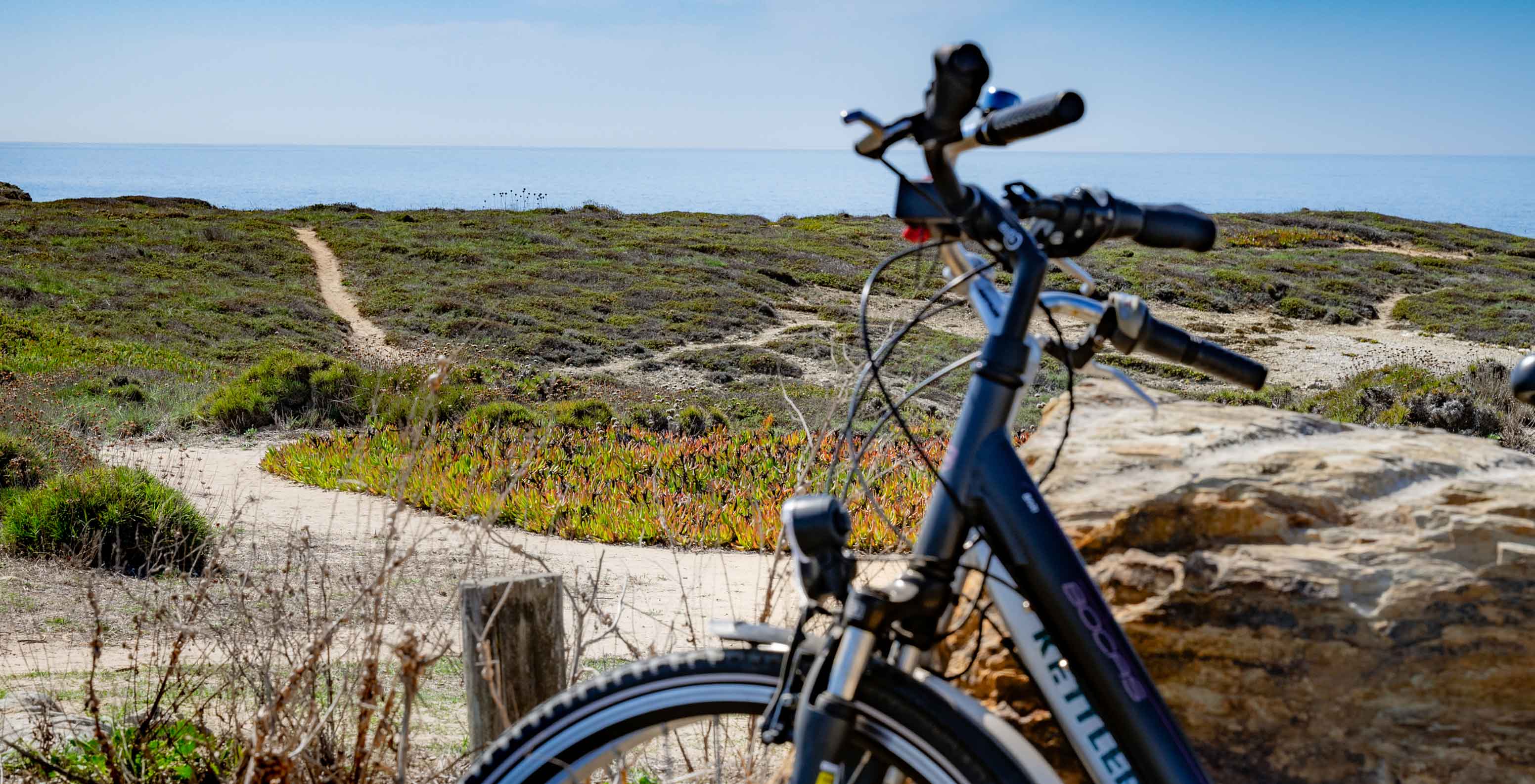 Bicicleta junto a trilho de areia e vegetação costeira com vista para o mar no Pestana Porto Covo Village