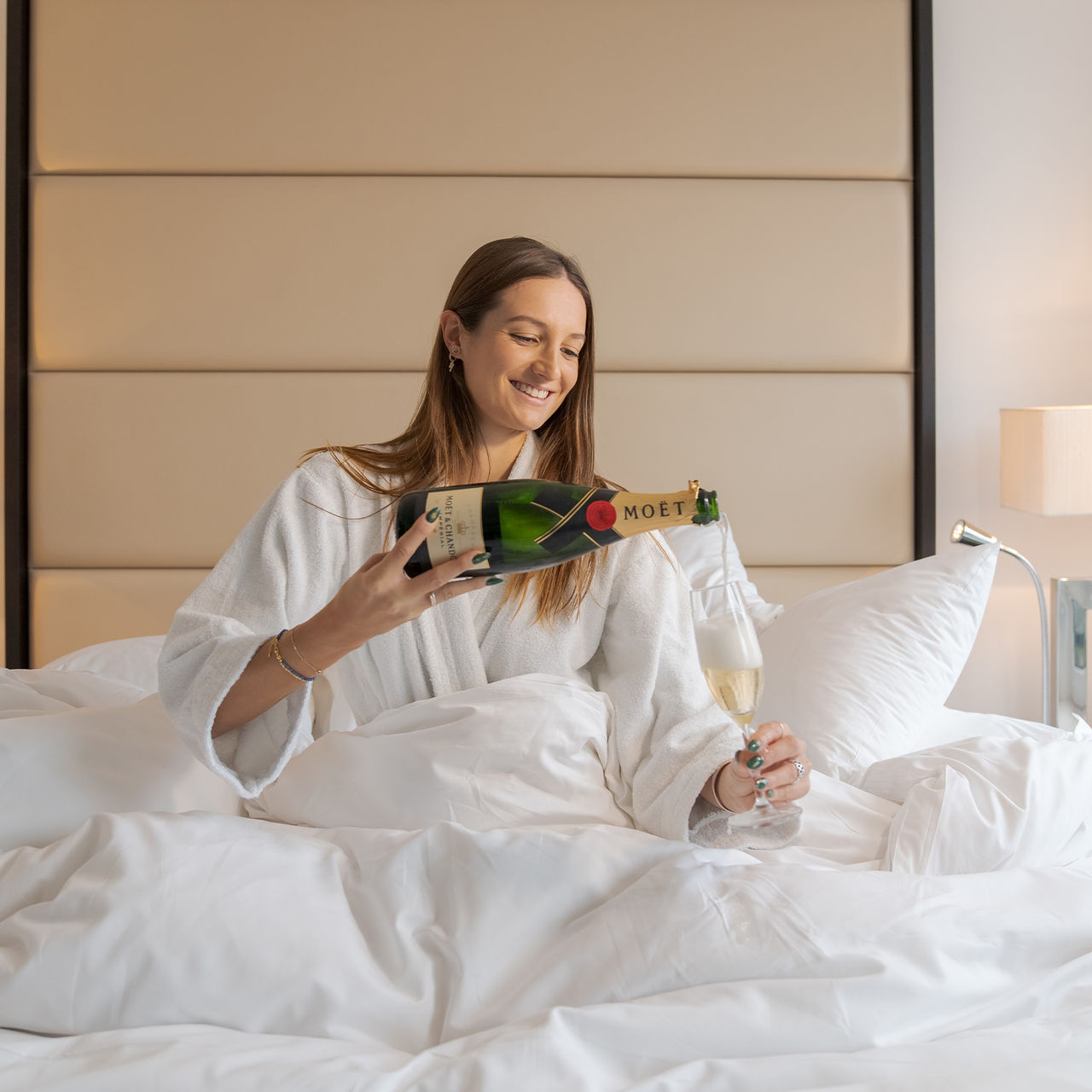 Vrouw lachend op bed met witte badjas en champagne