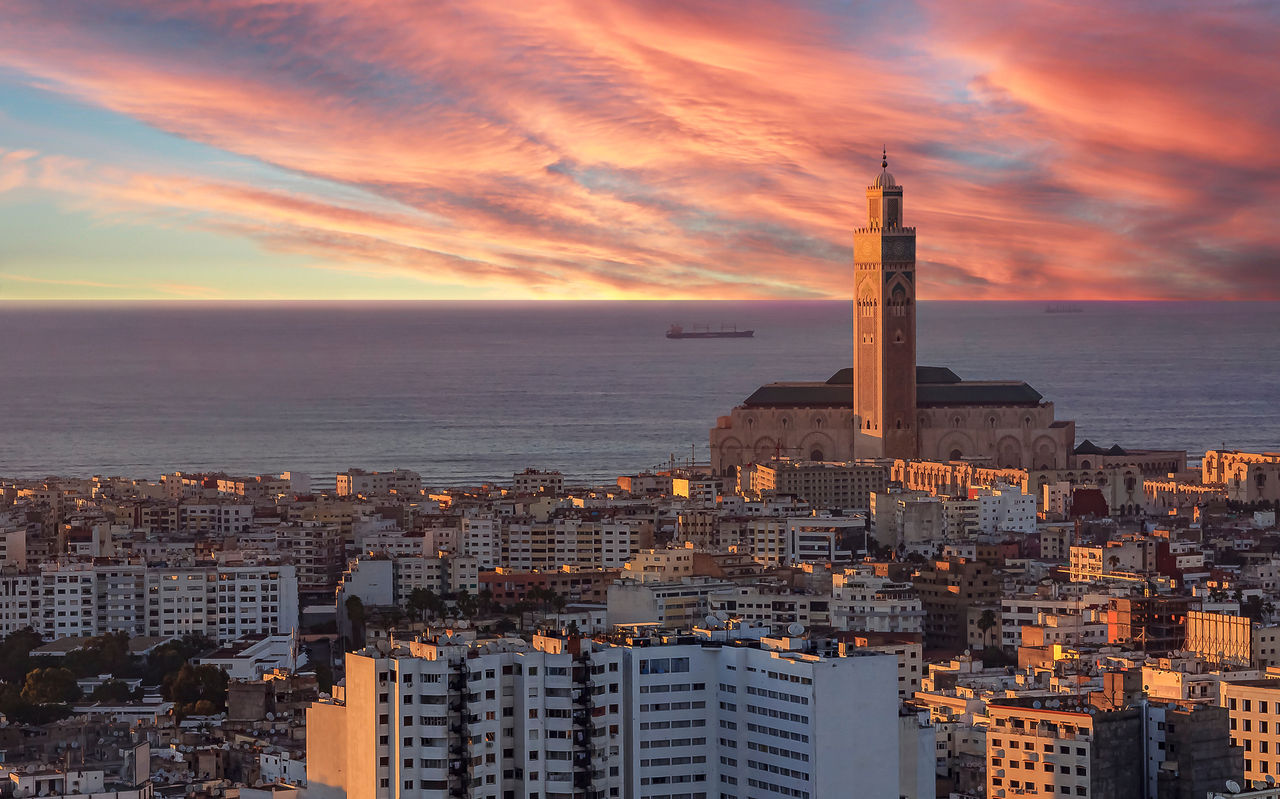 Aerial view over Casablanca, with the sunset illuminating various buildings, the sky over the sea, and the tower