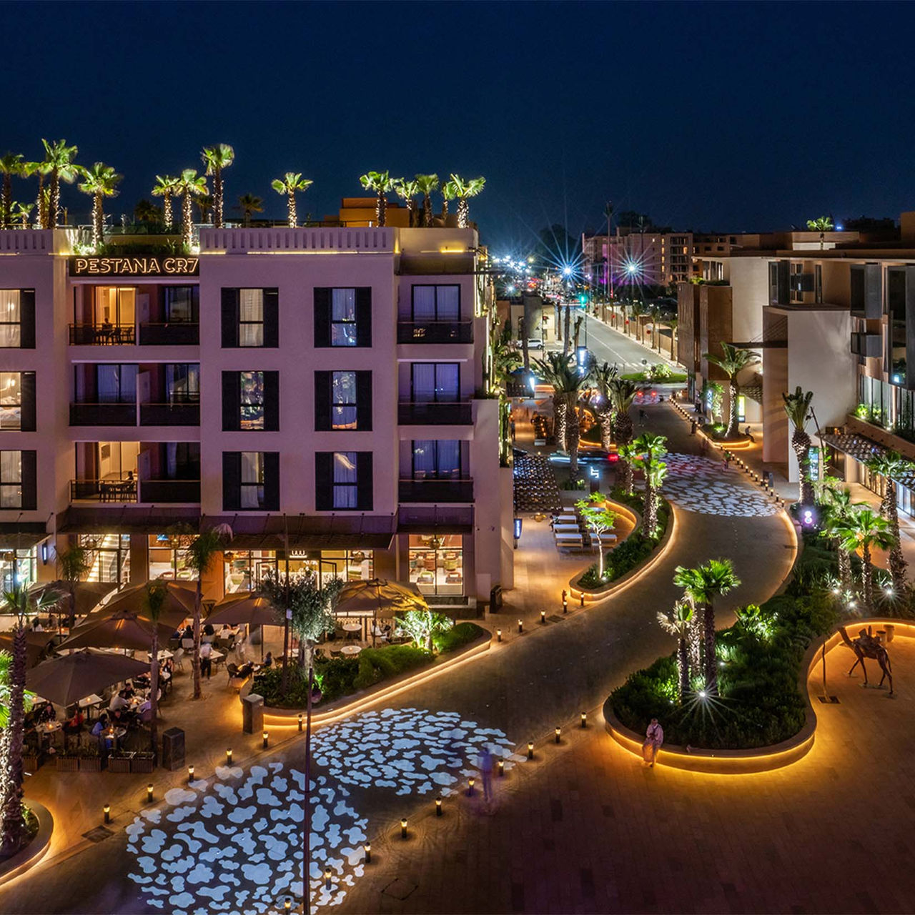 Exterior at night of Pestana CR7 Marrakech, located on M Avenue, with several shops, palm trees, and lit shrubs