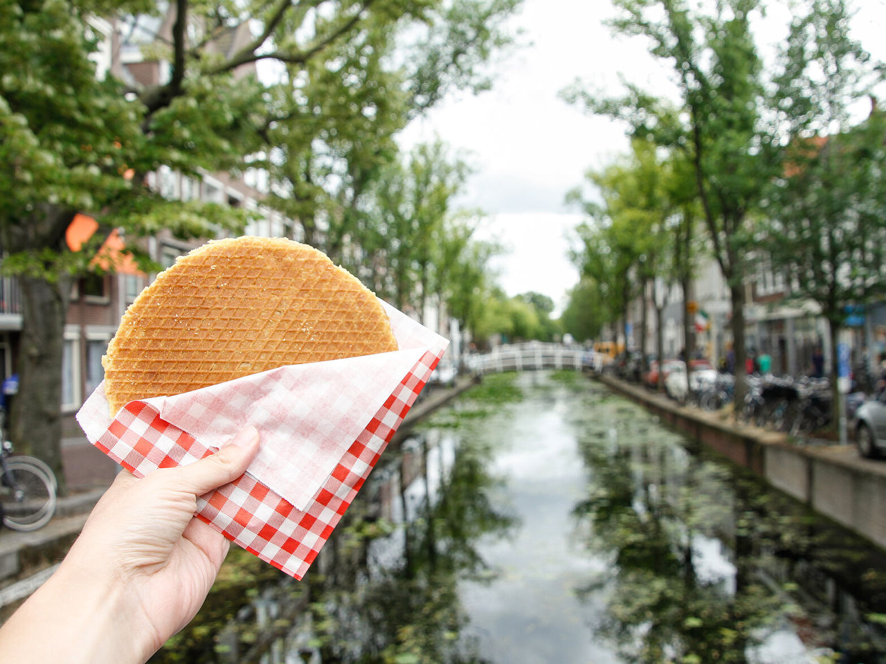 Hand holding a Dutch cookie, known as Stroopwafel, filled with caramel, with a canal in the background