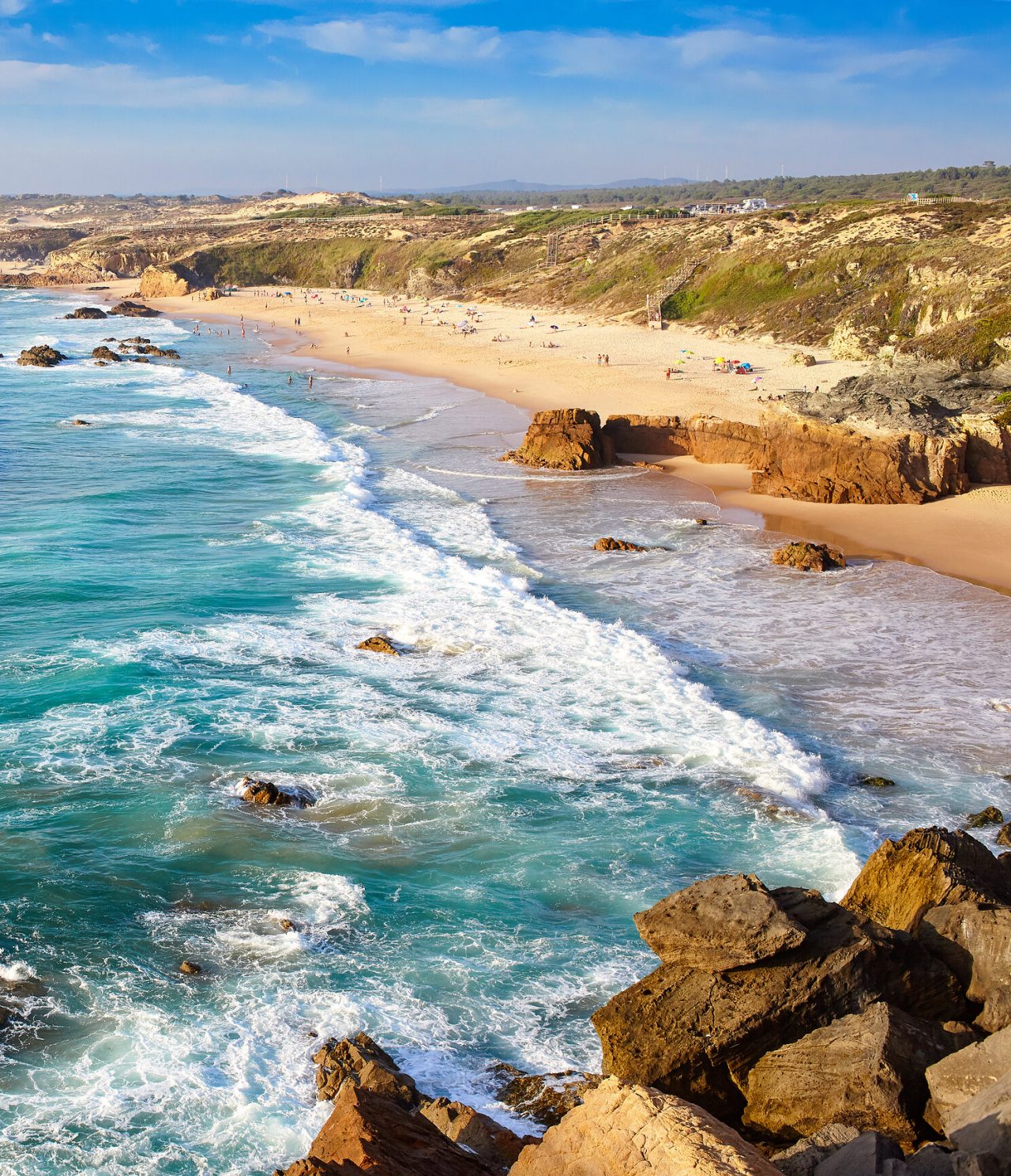 Panoramic view of the Alentejo coast, with a vast golden sandy beach, rocks and rough sea, under sunlight