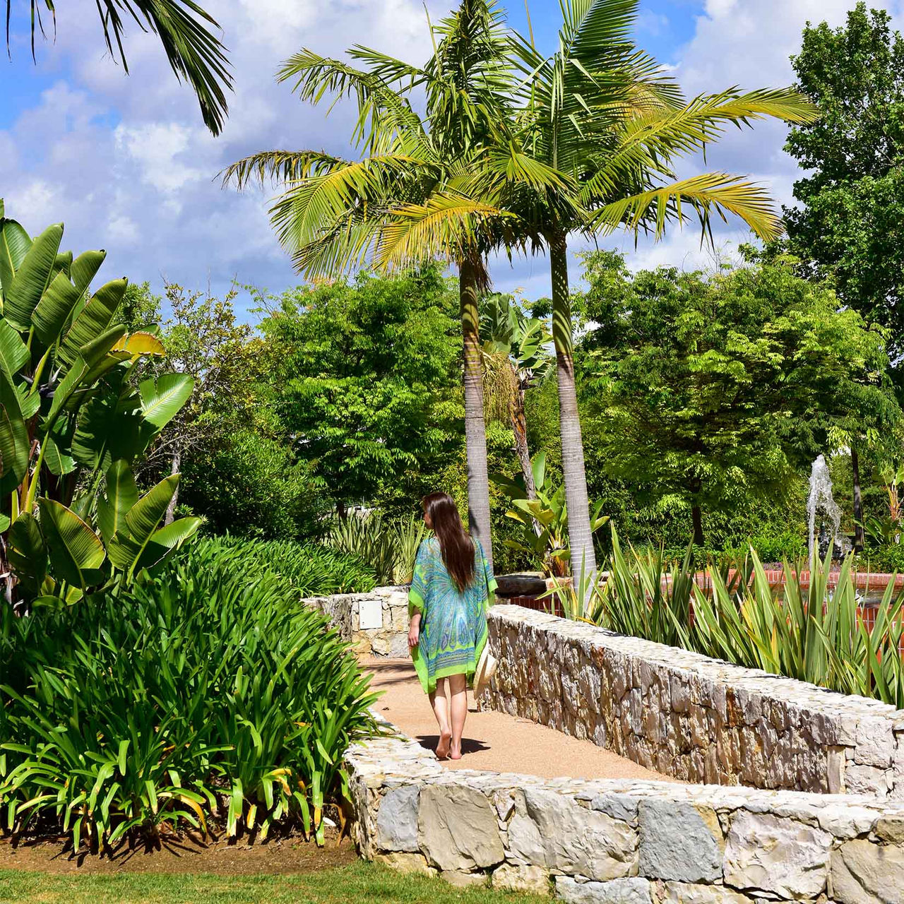 Garden with grass, palm trees, and various trees, with a lady on the walkway with stone walls
