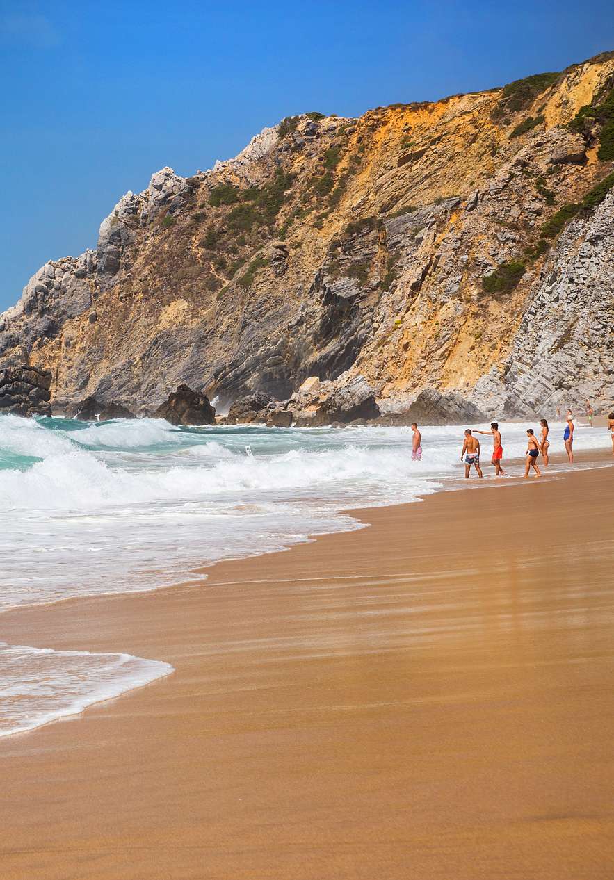Beach in Sintra with golden sand and waves breaking on the shore with people entering the water and rocks in the background