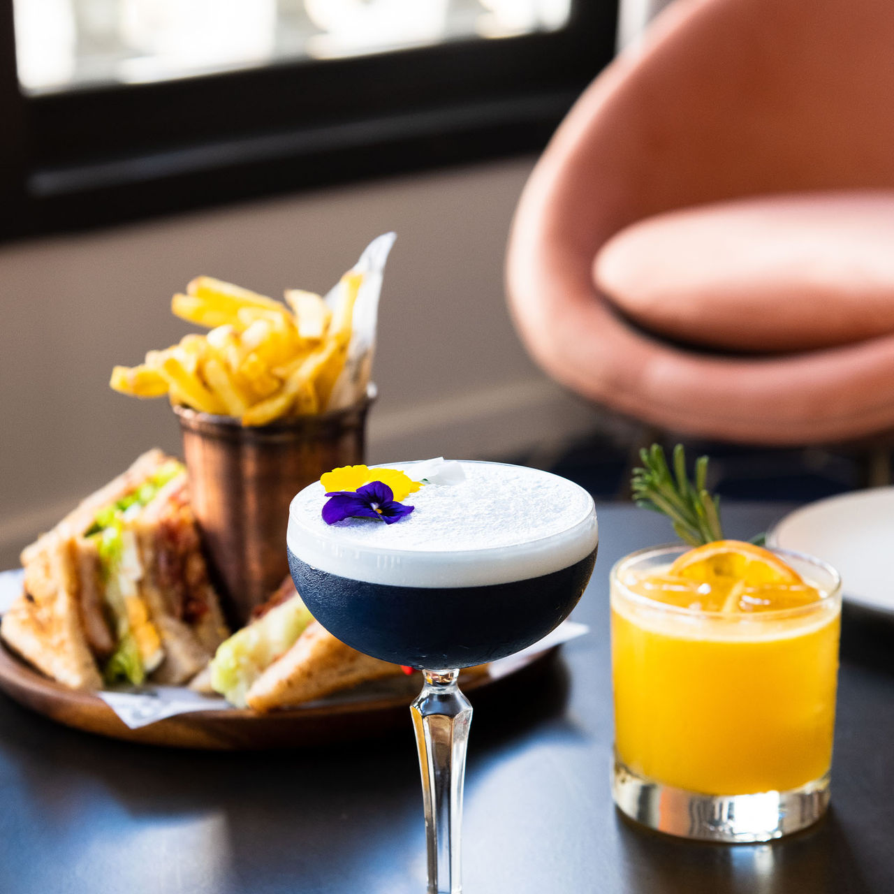 Side view of a cocktail decorated with petals, an orange drink with herbs, and a toast with fries